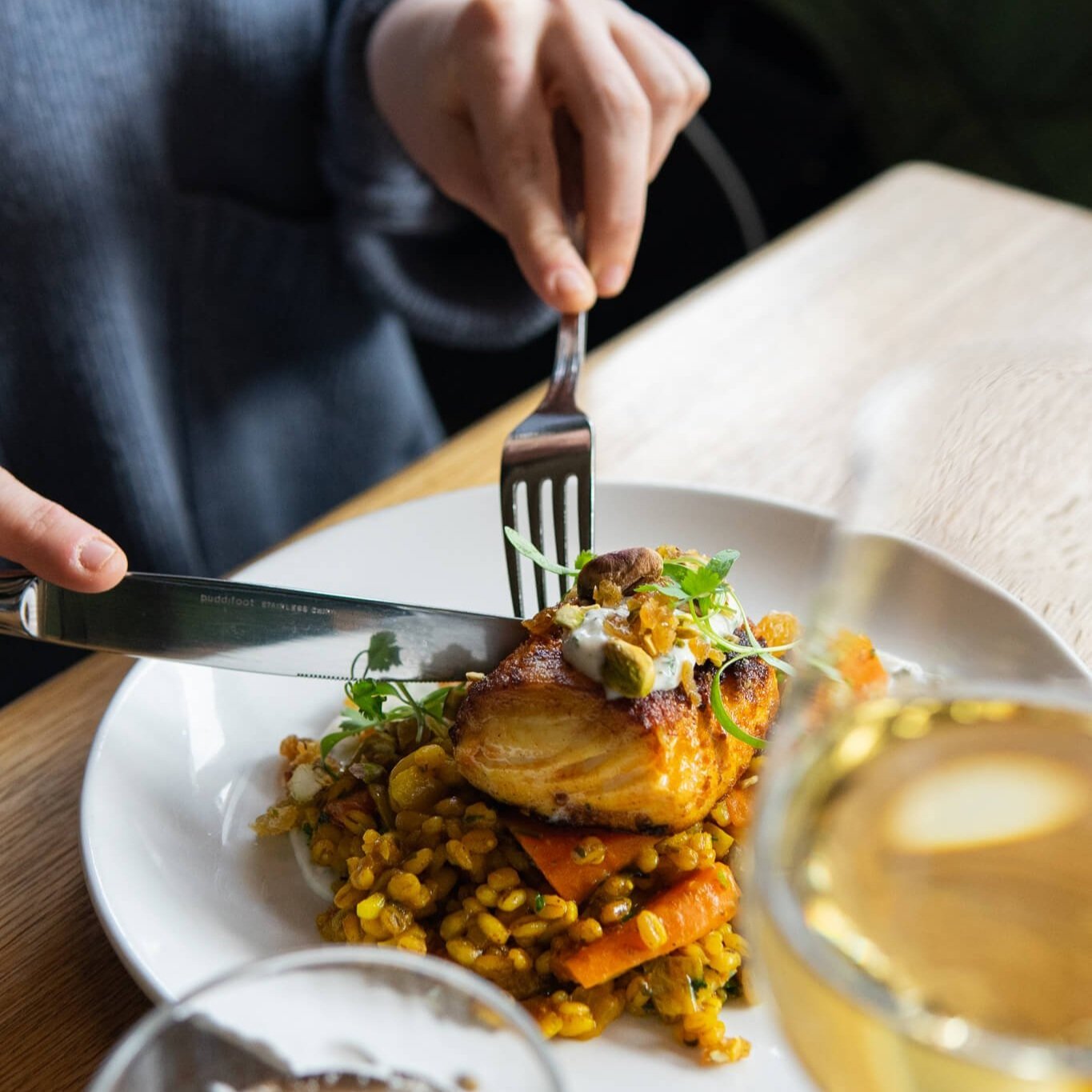 A close up photo of a customer's plate of food as they cut into it for the first bite. Out of focus are wine glasses. The food piled on the plate looks like a grilled fish piled on top of a mix of grains and vegetables, beautifully garnished on a whi