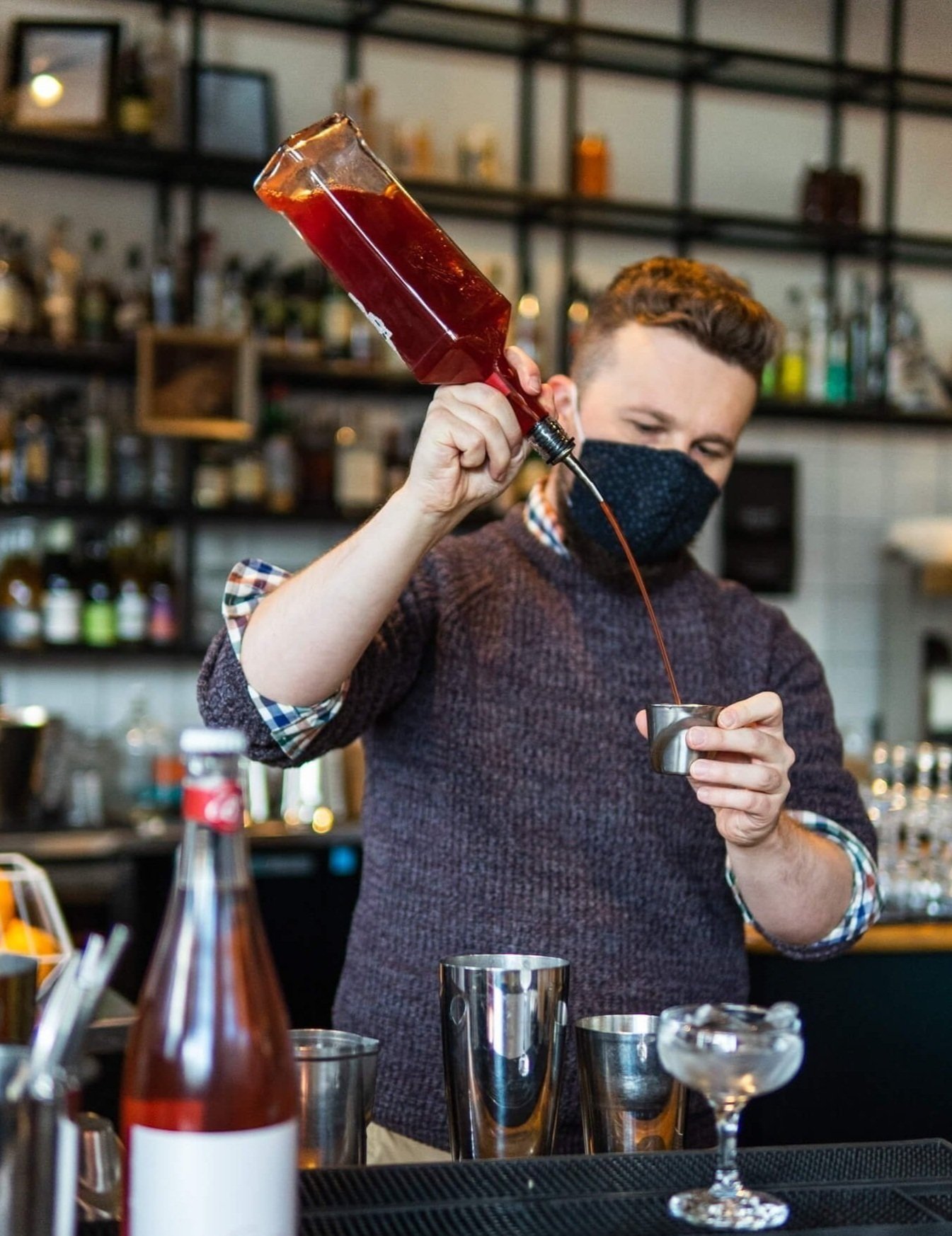 Shot from the perspective of a person sitting at a bar, a bartender is behind the bar pouring a red liquid into a shot glass. Around him there are glasses, a metal shaker, and liquor bottles out of focus.