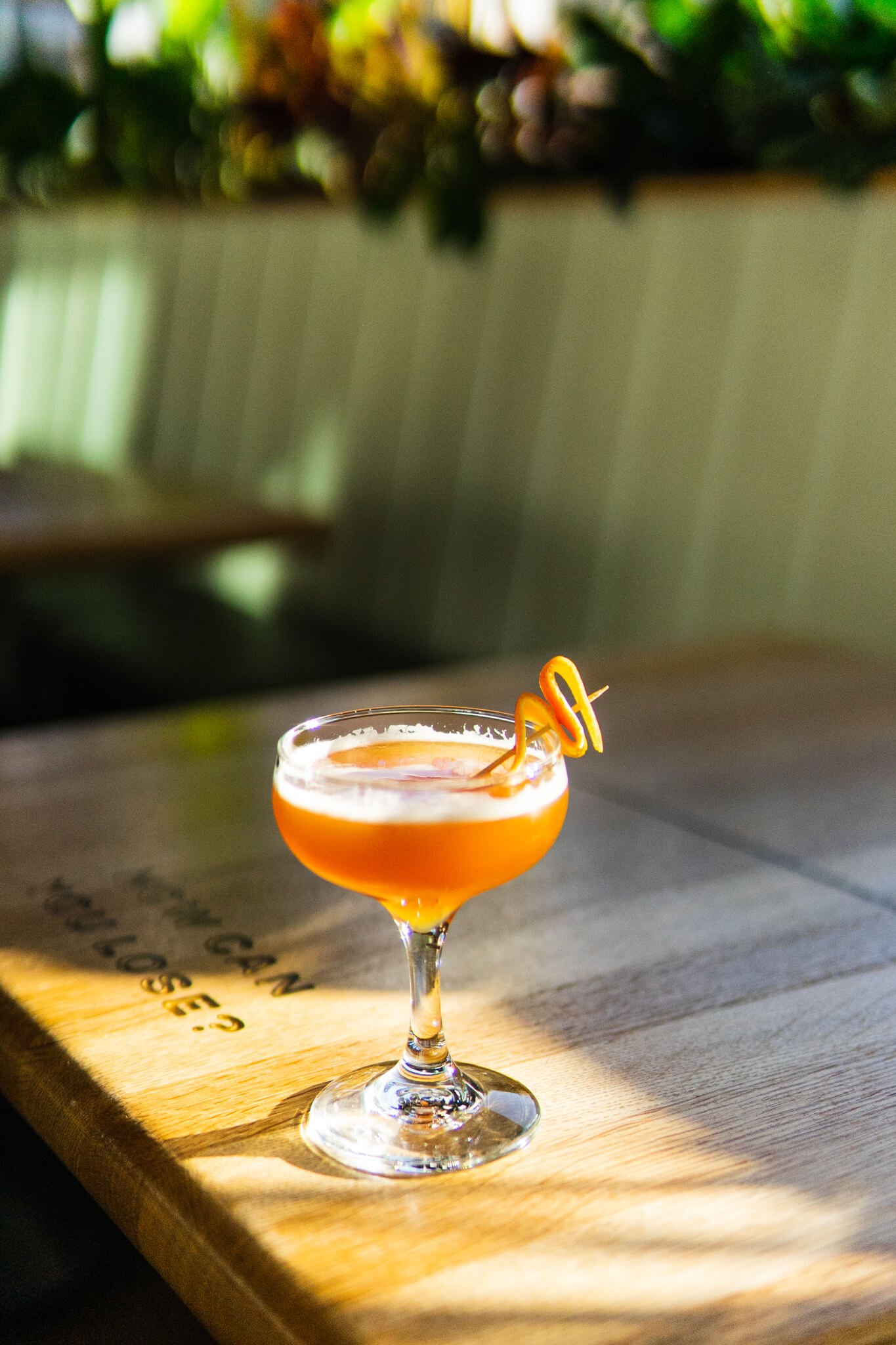 A beautiful orange cocktail in a coupe sits on a table in a sunlit spot. It is garnished with a orange twist and appears effervescent.