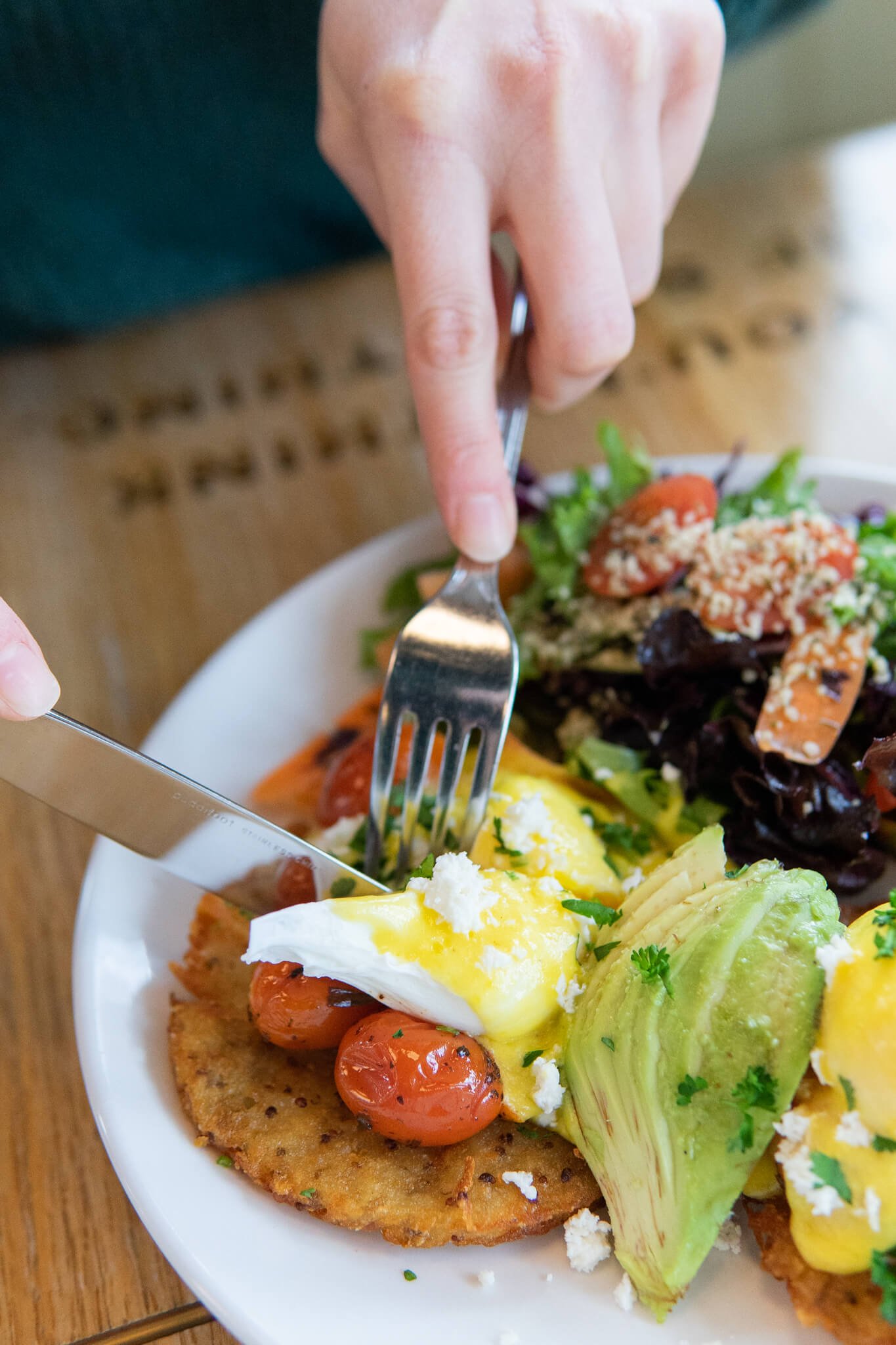 A close up photo of an egg on a hashbrown patty, blistered cherry tomatoes, avocado and a side salad. They are covered in a herb blend and a customer's hands are in view holding their fork and knife while cutting into the egg and hashbrown.