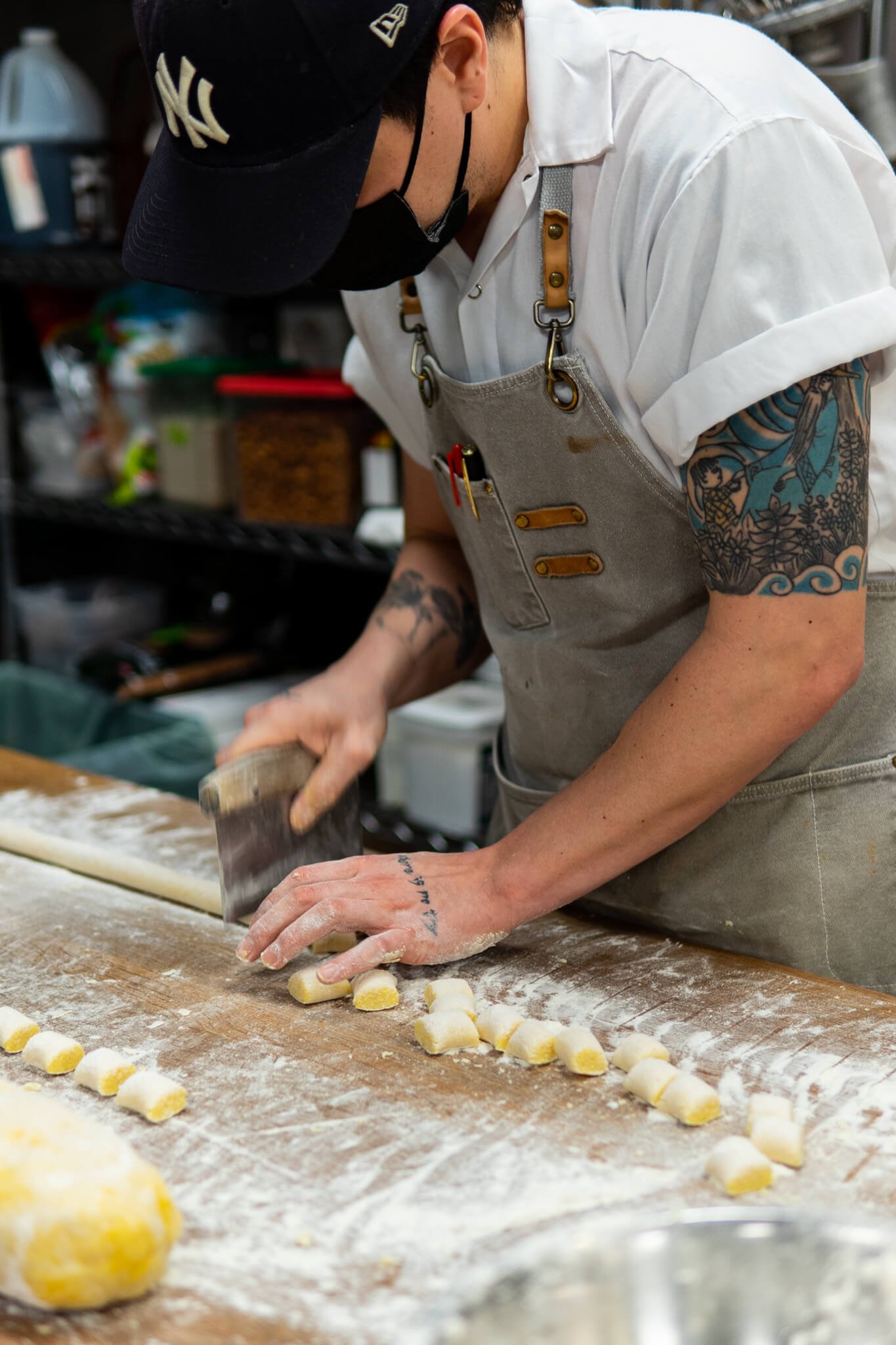 A cook from the kitchen is at a work table. He's wearing a hat, a kitchen apron, and a button up tshirt. He is working at a table covered in flour and is cutting up a long tube of dough appearing to make gnocchi