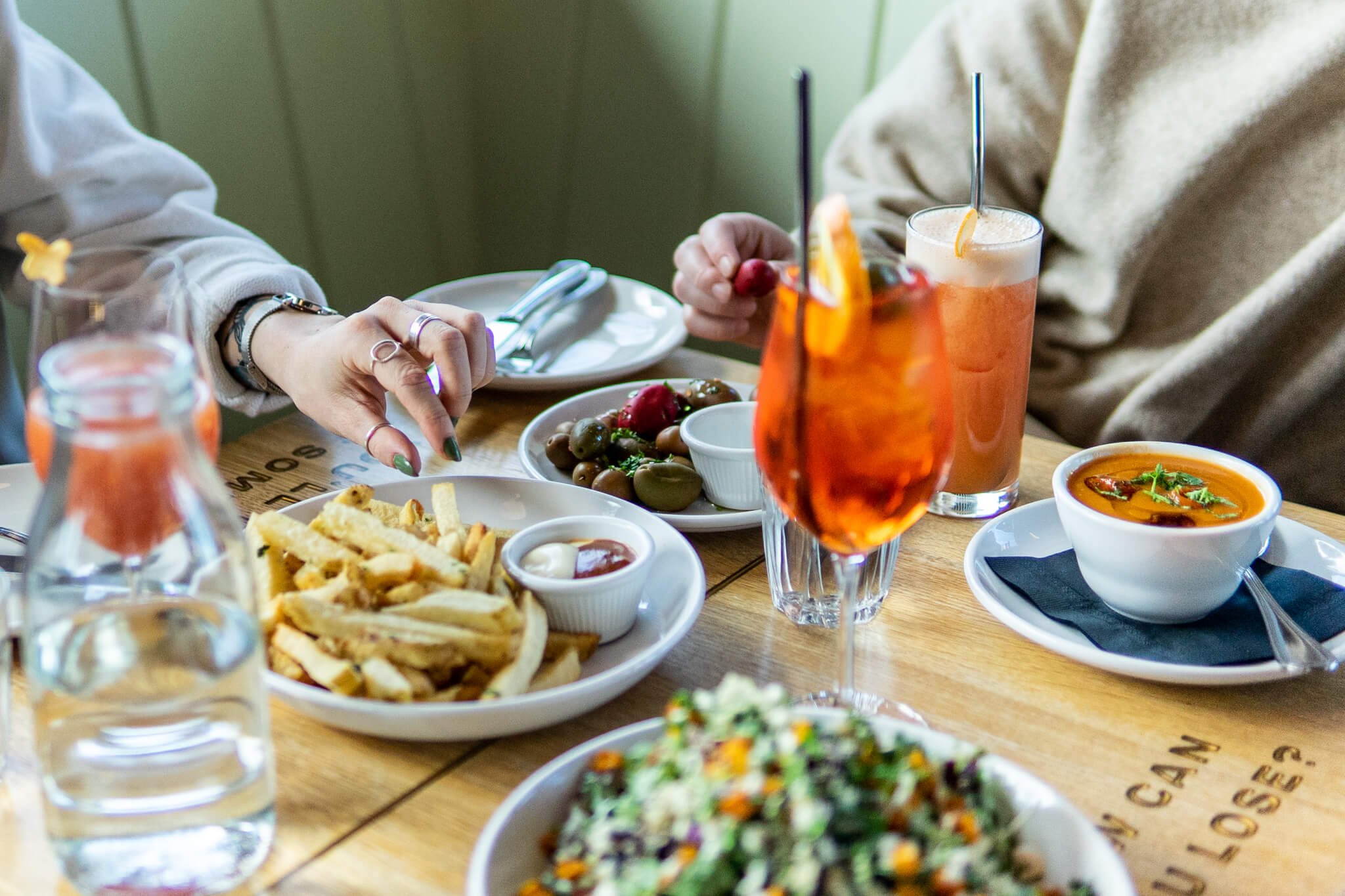 A table full of various different share plates are in view including salad, bowl of soup, brussel sprouts, and french fries as well as cocktails. There are two people sitting at the table, just their hands are visible. A hand is moving into frame abo
